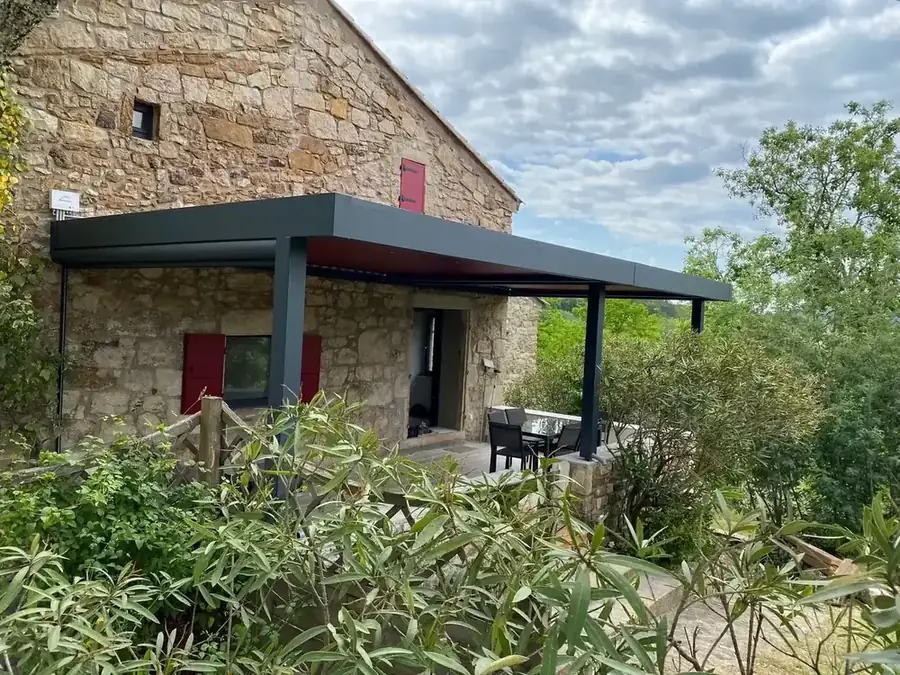 Pergola anthracite en façade d'une maison en pierre aux volets rouges