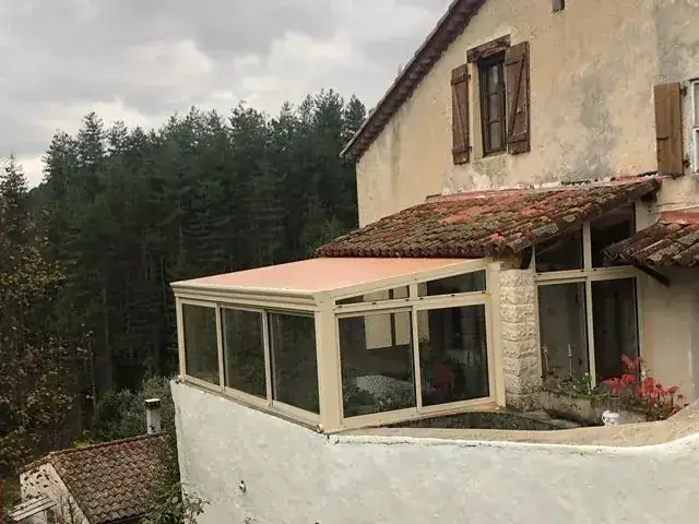 Véranda beige accrochée à une maison en pierre ancienne, forêt en arrière-plan